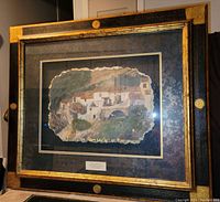 Full view of the large professionally framed watercolor painting on plaster showing rustic houses and a bridge, with a patterned dark mat and gold black frame.