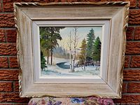Framed oil painting showing winter river landscape with trees and snow, signature visible, photographed against a brick wall.