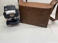 Polaroid SX-70 Land Camera Sonar OneStep with detachable flash in front of brown Polaroid branded leather carrying case