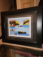 Image of framed Maud Lewis print showing colorful coastal scene with boats, dock, seagulls, and houses surrounded by autumn trees. Visible signature and title on print.