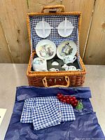 Small wicker picnic basket open showing miniature Reutter Porcelain teaset with Peter Rabbit illustrations inside, plus blue gingham cloth and napkins and artificial grapes outside the basket.