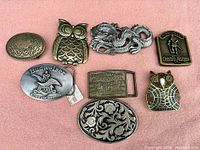 Full view of all eight vintage belt buckles arranged on pink cloth surface showing condition and design details