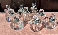 Group of 10 small vintage cut crystal perfume bottles with faceted round tops and glass applicators in lids, arranged on a pink surface with dark background, showing clear crystal shine and cuts.