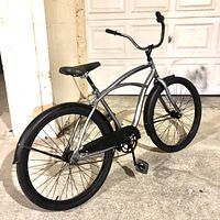Rear-angled view of the gray beach cruiser bicycle showing frame, rear wheel, fender, saddle, pedals, chain guard, and handlebars.
