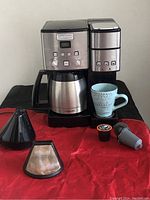Full view of Cuisinart Coffee Center showing 12 cup coffeemaker with thermal carafe, single-serve brewer, ceramic mug, reusable filter, paper filter, and two K-Cup pods on red cloth.