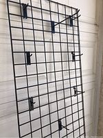 Full view of black metal grid wall panel with six attached hooks displayed vertically against a white background.