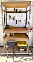 Front view of shelving unit showing four wooden shelves and pegboard back. Stool and wheeled base also visible. Shelves contain a few small items and tools.