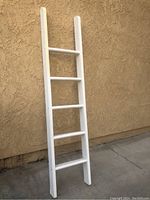 White wooden ladder with five rungs leaning against a beige stucco wall on concrete ground.