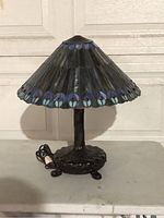 Photo of Tiffany style lamp showing stained glass shade with dark panels and peacock feather pattern edge, metal base and cord attached.