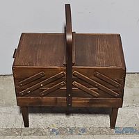 Front view of closed teak wood accordion style sewing basket with central handle and four legs.