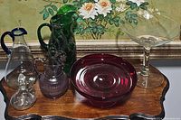 Set of six glass serving pieces displayed on a wooden surface in front of floral artwork, includes clear pedestal dish, red pedestal server, green floral pitcher, purple pitcher, hand-blown pitcher with blue trim, and cruet.