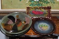 Photo of three serving dishes together on wood surface with floral painting in background: one large oval Lucite bowl with abstract pattern, rectangular ceramic tray with multicolor design, round ceramic plate with leaf pattern.