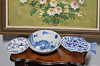 Three blue and white china pieces displayed on wooden table beneath floral painting; includes Winterthur dragon bowl, fish-shaped plate, Liesbeek Studio round plate