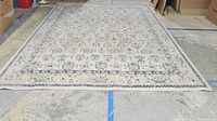 Full view of the beige Brielle rug with intricate patterns laid out on a concrete floor in a warehouse setting.