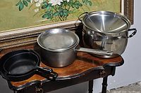 Two cast iron pans, stainless steel saucepan with lid, and stainless steel stock pot with lid on a wooden table with floral painting in background.