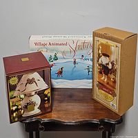 Three boxed Christmas decor items on a wooden table: Village Animated Skating Pond, wooden Santa nutcracker in box, and a holiday tartan candle lamp in a box.