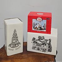 Three boxes of Christmas village collectibles arranged on a wooden table, showing packaging with images of village houses and Christmas tree.