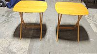 Two wooden folding TV trays standing side by side on concrete floor, showing full trays and legs.