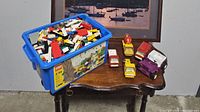 Blue Lego Classic container filled with assorted loose Lego bricks beside five vintage toy trucks on a wooden table.