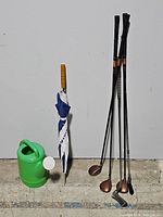 Photo of 4 golf clubs, a green water can, and a blue/white closed umbrella leaning against a white wall