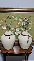 Two cream ceramic table lamps side by side on a wooden table with floral painting background.