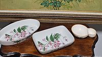 Three dishes on a wooden surface. Two oval dishes with pink and purple flowers and one small two compartment olive dish with blue and brown floral painting.