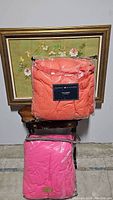 Two Tommy Hilfiger comforters stacked, one coral orange and one bright pink, both full/queen size, in clear plastic packaging.