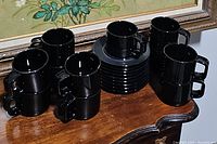 Side view of stacked black cups and grouped saucers on a wooden table under a framed floral artwork.