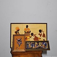 Three framed art pieces shown together on a wooden table against a gray wall. One large fabric art with African figures and two smaller yarn art pieces, one vertical and one horizontal, are visible. One frame shows wear and damage.