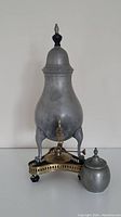 Frontal view of antique pewter tap jug on brass and black wooden stand with brass oil lamp and pewter lidded container in front