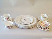 Photo showing assorted tea cups with matching saucers, bowls, and plates arranged on flat surface. Floral patterns and delicate china/porcelain pieces.