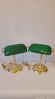 Pair of green glass shade desk lamps with brass bases, pull chains and cords, positioned side by side.