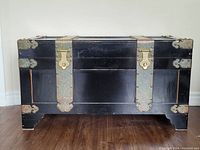 Front view of large black wooden trunk with brass straps and corner protectors.
