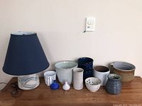 Photo showing entire lot: ceramic lamp with navy shade, assorted pottery pots and vases of various sizes and colors arranged on wooden table against beige wall.