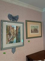 Two framed art prints hung on a pink wall. The left one is a colorful Venetian canal scene with boats, framed in grey, topped with a fabric bow. The right one is a sepia-toned architectural print in a green frame.