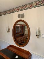Wall with an oval mirror in cherry wood frame accompanied by two brass sconces with white candles and glass globes, items mounted on wall above wooden surface.