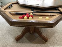 Detailed view of the bumper pool table showing the octagonal shape, felt surface, two pool cues, red and yellow balls, and wooden base with paint splatters.