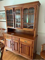 Front view of two-piece cherry wood hutch with glass upper doors and wooden lower doors and drawers, wooden dining chairs next to it.