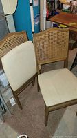 Two wooden folding chairs with cane backs and cream vinyl seats shown side by side on carpet in a basement or utility area. Both chairs have visible stains and discoloration on seats.