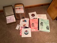 Full view of lot showing multiple 45 rpm records in paper sleeves and a box plus several jukebox manuals spread on carpeted floor.
