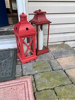Two red metal framed lanterns with glass panels and white faux candles inside, placed on stone flooring beside a door.