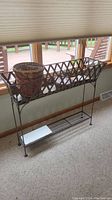 Side and front view of black metal basket style plant stand with worn woven basket planter inside, placed indoors near window.