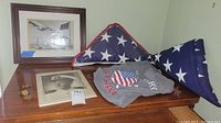 Photo of two framed photos, two folded American flags in display cases, grey veteran shirt, and ribbon medal on wooden surface