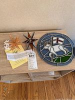 Photo showing three wooden Moravian stars of different sizes and colors alongside a round stained glass panel with a lamb and flag design on a wooden surface.
