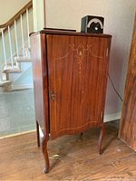 Front and side views of a wooden music cabinet showing decorative inlay on the front door, key lock, and cabriole legs.