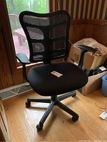 Front and angled views of a black office chair with mesh back, padded seat, armrests, and wheels on hardwood floor near window.