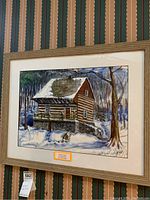 Frontal image of the painting showing the snow-covered cabin in a wooded environment with the frame and matting intact.