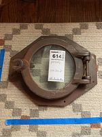 Front view of metal porthole window showing circular glass and latch mechanism placed on carpet with label.