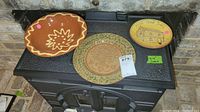 Three redware plates displayed on a black surface. Left plate is oval with scalloped edges and cream floral design, middle plate is round with green border and central inscription, and right plate is round with yellow base and cabin drawing with text.