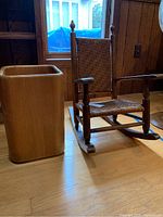 Photo showing both the child's rocking chair with woven back and seat beside the wooden trash can on a wooden floor near a window.